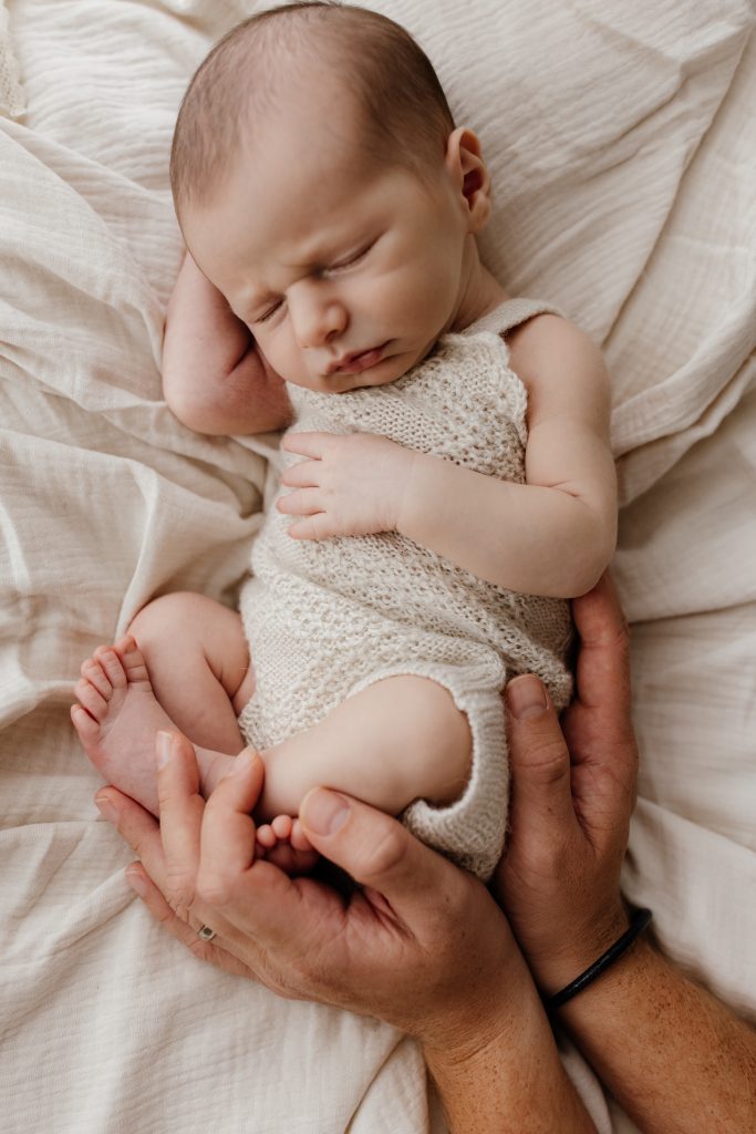 A newborn baby curled up with mums hands holding on to him. Wearing a plain cream onesie.