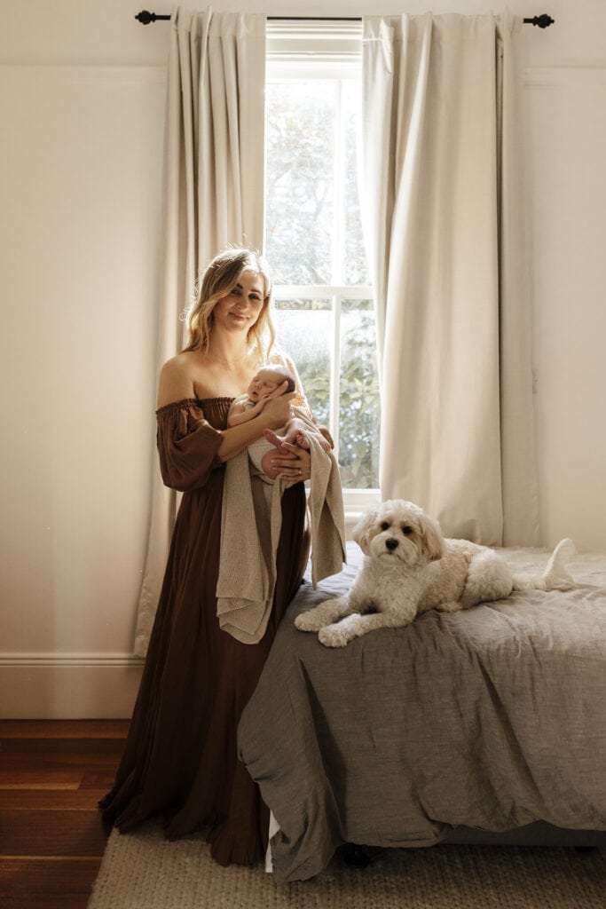 A mother holding her new baby in a sunlit room with her dog.