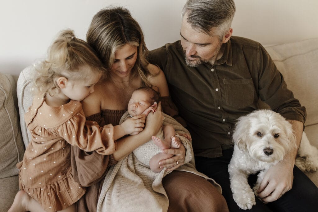 A family of four and their dog cuddles on the couch for their newborn photoshoot. Wearing warm soft colours.