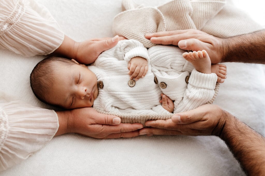 A newborn baby curled up in a white knitted onesie in his parents hands.
