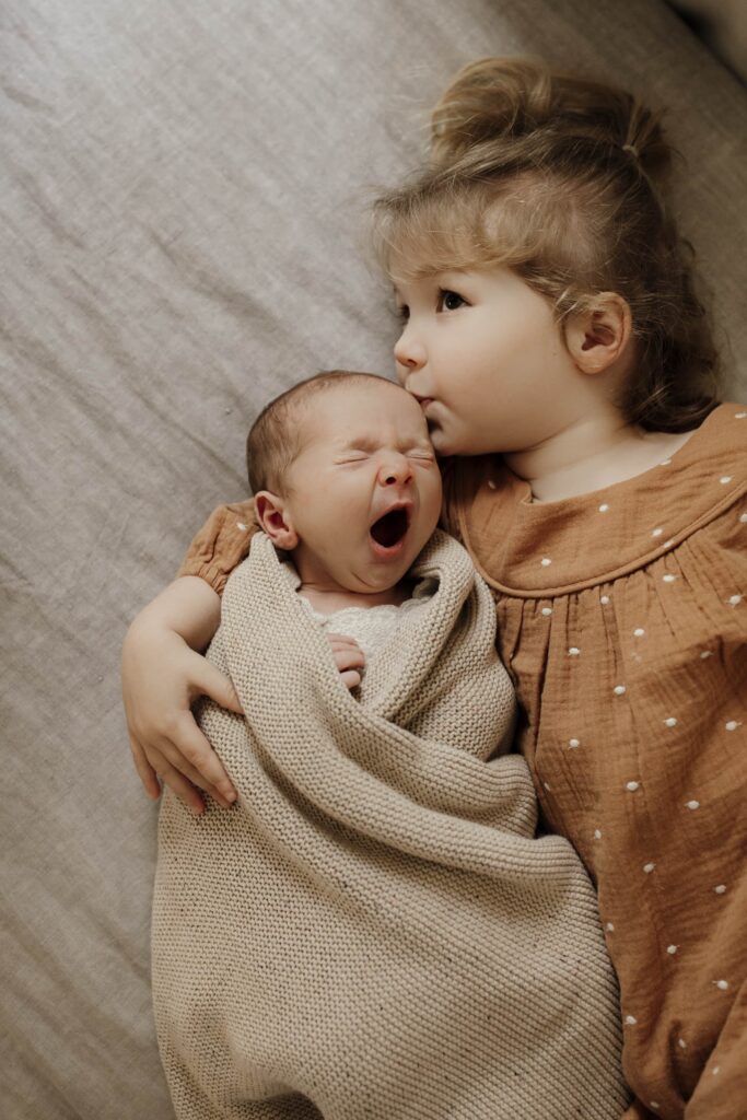 A newborn baby curled up in his big sisters arms while he yawns himself to sleep.
