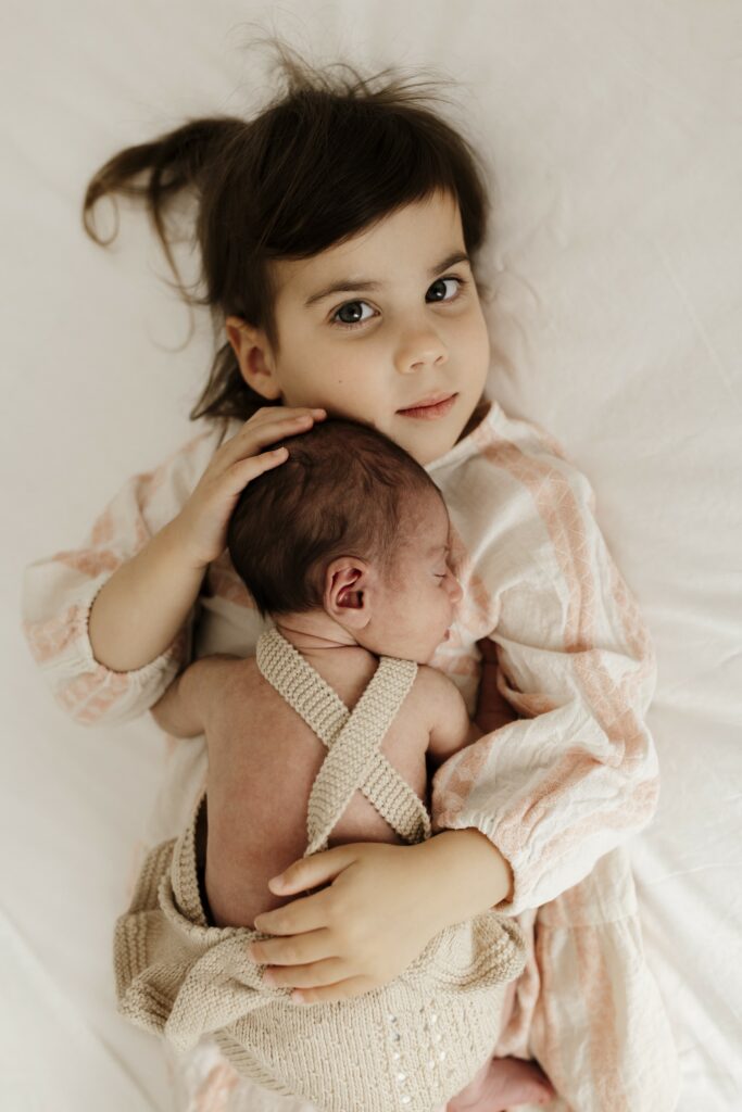 A 3 year old girl holding her new baby sister while she sleeps on her chest. 